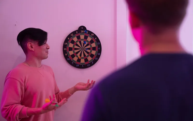 A man holding darts stands by a dartboard, laughing at his opponent
