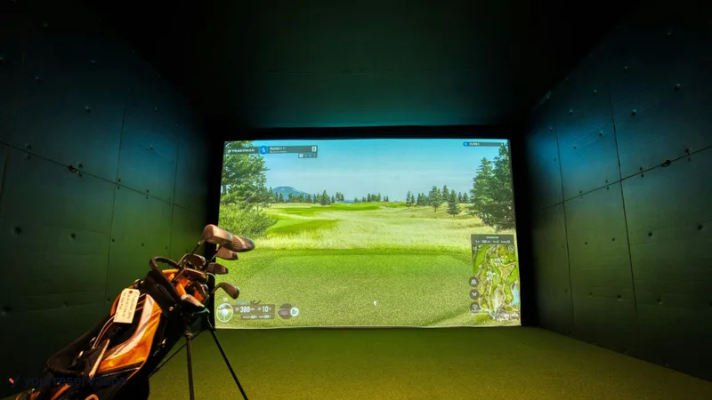 A golf bag stands beside the hitting mat in a dark simulator bay with a golf course on the screen