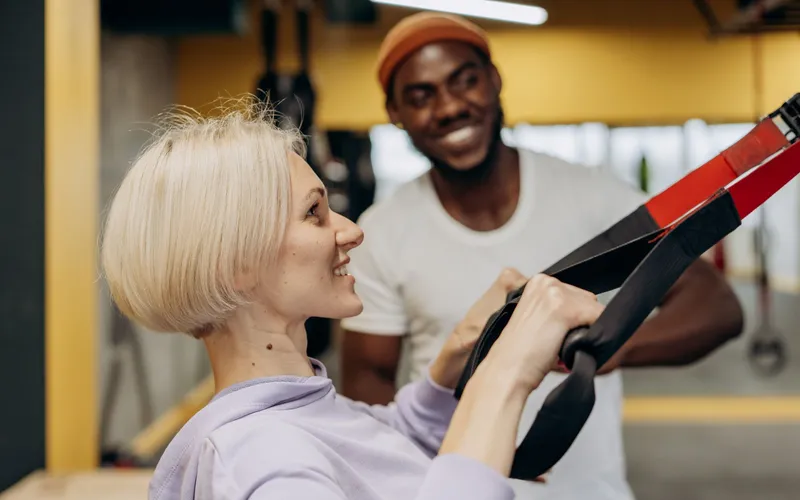 Ein Personal Trainer schaut lächelnd zu, während eine Kundin mit Schlingentrainer übt