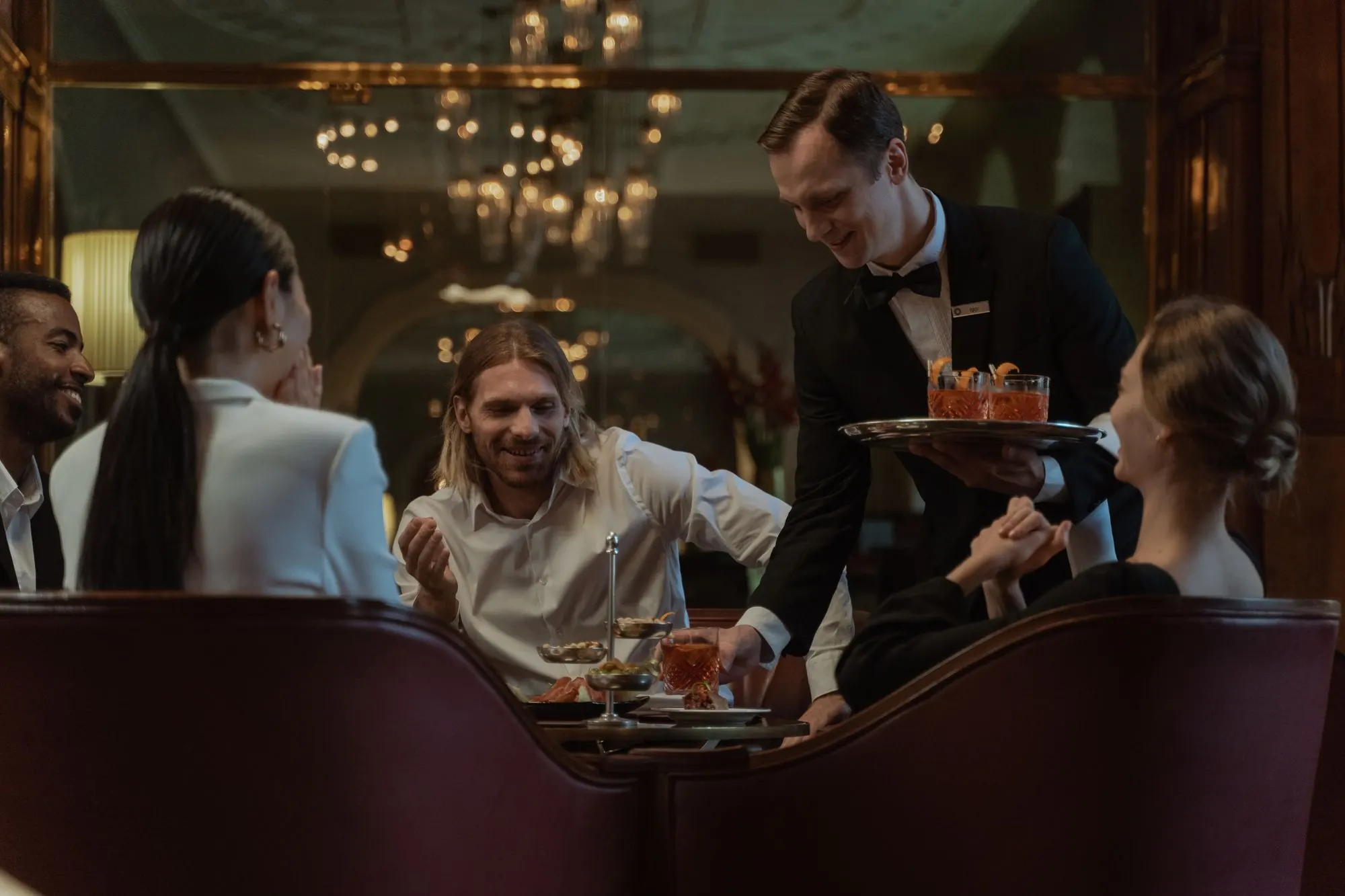 En servitör serverar drinkar till ett bord med fyra skrattande gäster i en varmt upplyst restaurang