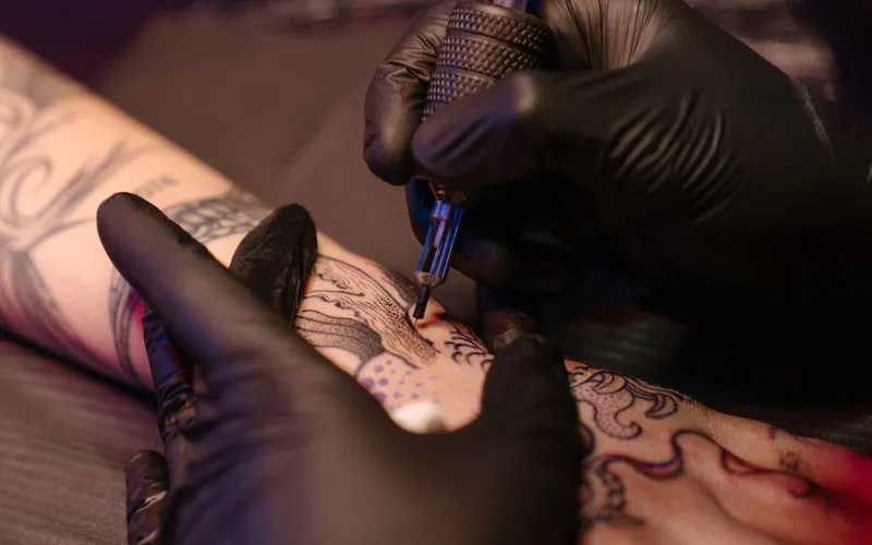 Hands in black gloves tattooing a client's hand with a tattoo machine