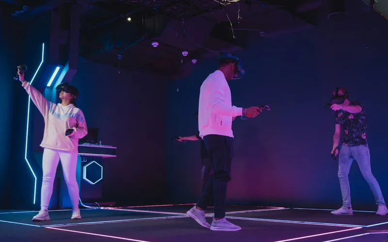 Three players wearing VR headsets and holding controllers move around a dark play space with light strips on the floor